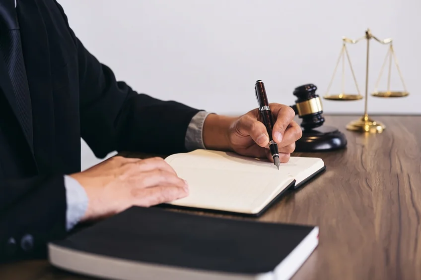 A person signing documents beside a gavel and the scales of justice.